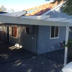House with grey corrugated cladding, tiled driveway, and white pergola.