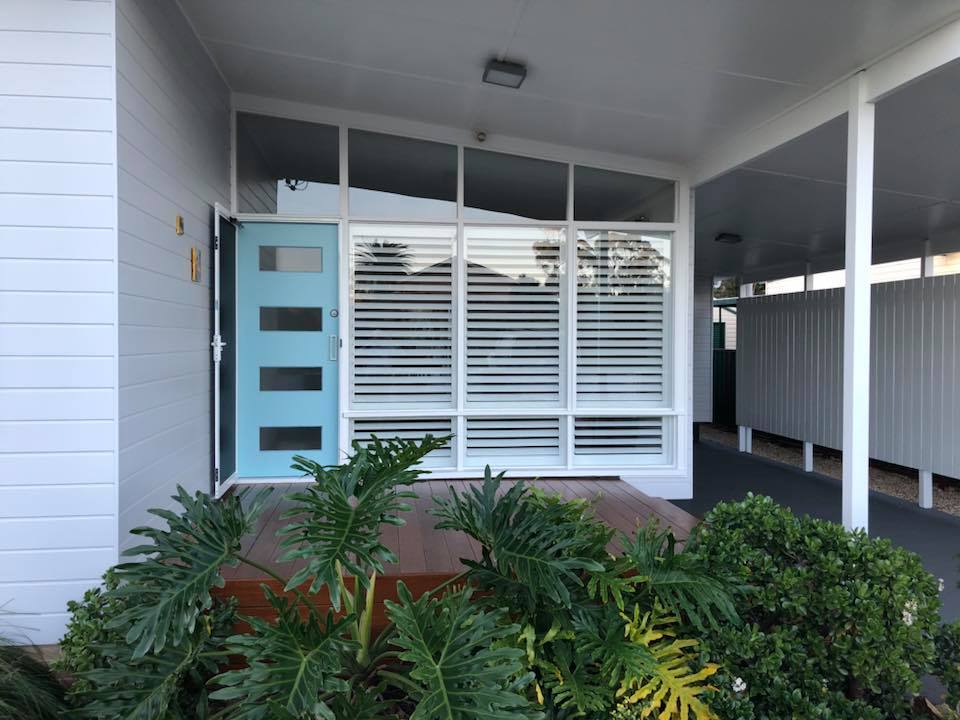 Front entrance with blue door, large windows with white shutters, and greenery.